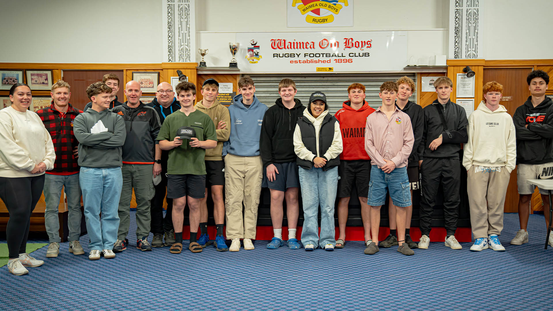 Group of young men posing for a photo in a gymnasium with 'Waimea Boys Rugby Football Club' sign.